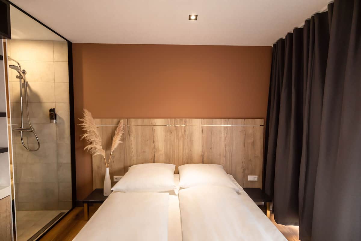 Ein modernes Hotelzimmer mit Dusche, einem Bett mit weißer Bettwäsche, einem Holz-Kopfende und einer Vase mit Pampasgras.