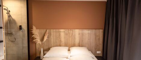 A modern hotel room with a shower, a bed with white linens, a wooden headboard, and a vase with pampas grass.