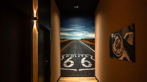 Un pasillo con suelo de madera que conduce a una carretera pintada en la pared, un cuadro enmarcado y una puerta a la izquierda.