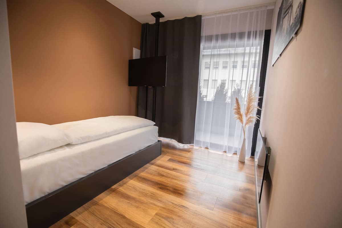 Ein modernes Schlafzimmer mit einem Bett, einem Flachbildfernseher an der Wand, einem Fenster mit leichtem Vorhang und Blick auf Gebäude.