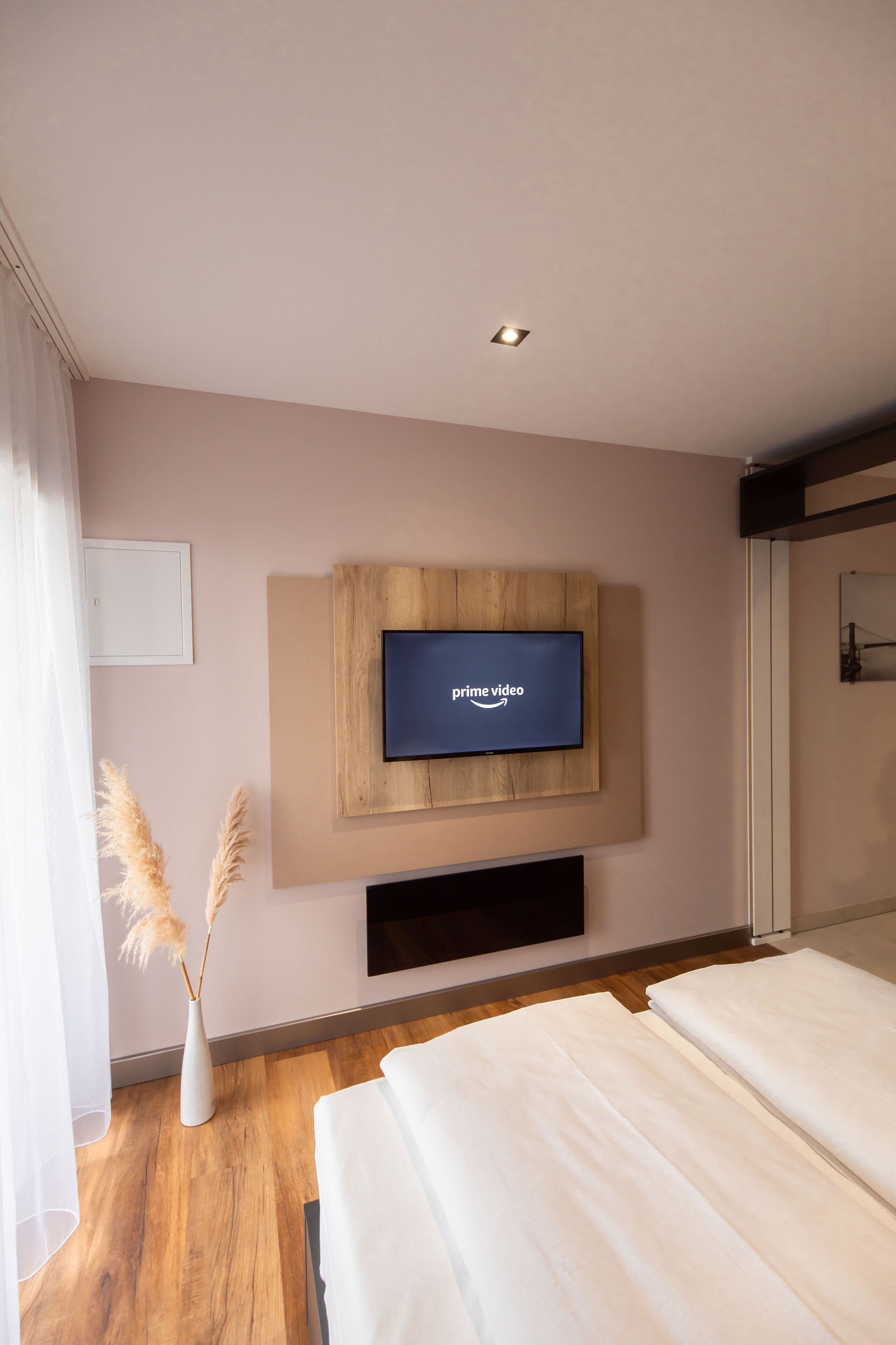 Una habitación de hotel moderna con un televisor de pantalla plana grande montado en un panel de madera, una cama blanca y piso de madera.