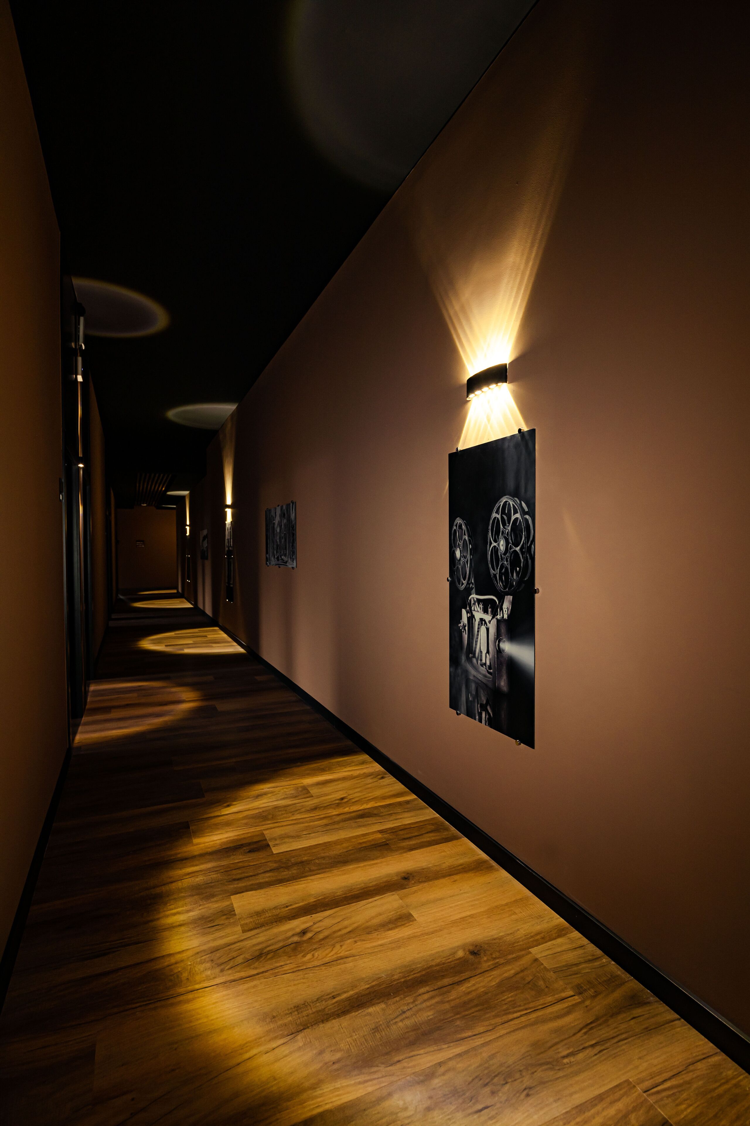 A hallway with wooden flooring, wall-mounted light fixtures, and framed artwork.