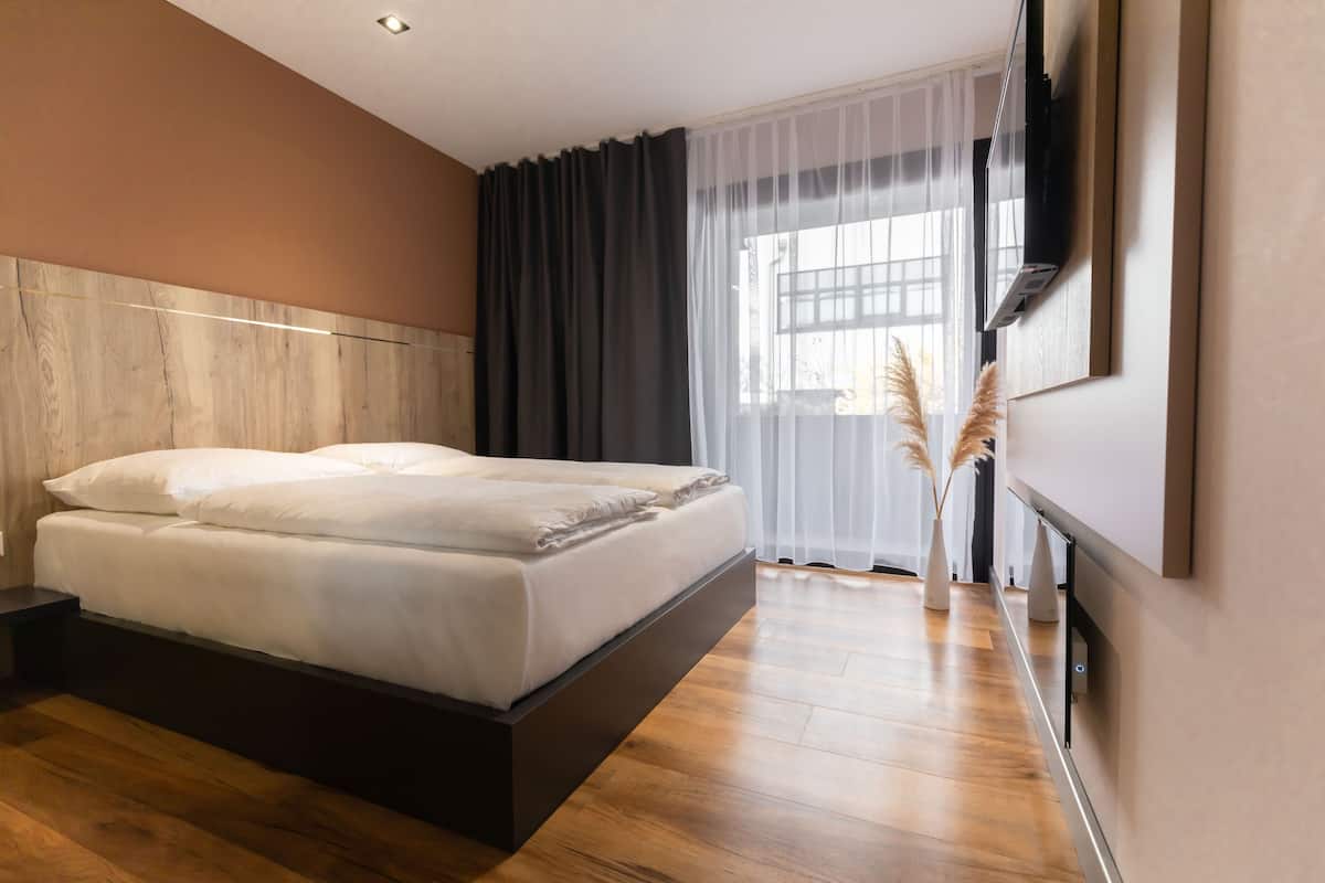 Ein modernes Schlafzimmer mit einem großen Bett, einem flachbildfernseher an der Wand und einem Fenster mit transparenten Vorhängen.