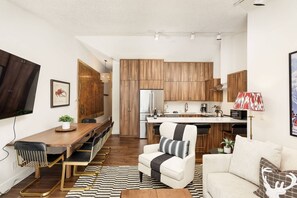 Una cocina moderna con armarios de madera, una encimera blanca y un horno empotrado. Hay un comedor con una mesa y sillas de madera, y un sofá en la sala de estar.