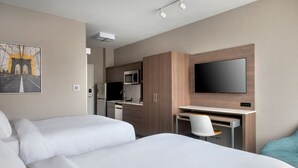 A hotel room with two beds, a desk with a chair, a flat-screen TV, a microwave, and a kitchen area.