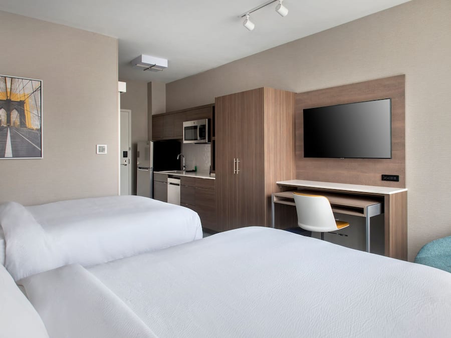 A hotel room with two beds, a desk with a chair, a flat-screen TV, a microwave, and a kitchen area.