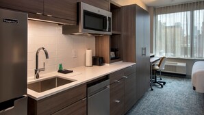 A modern kitchen with a built-in microwave, stainless steel sink, and dishwasher. There is a window with curtains and a bed in the adjacent room.