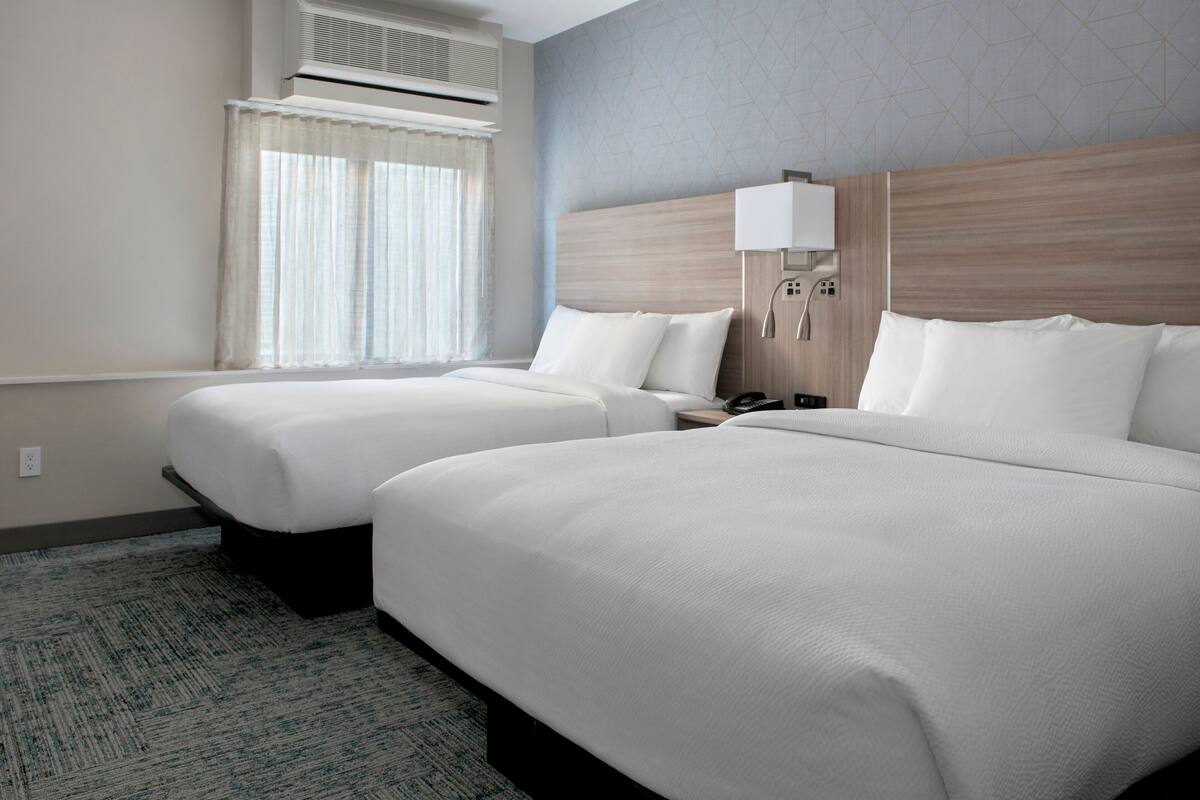 Habitació d'hotel amb dos llits, una finestra amb cortines, un aparell d'aire condicionat, una taula de nit amb una làmpada i un telèfon fixat a la paret.