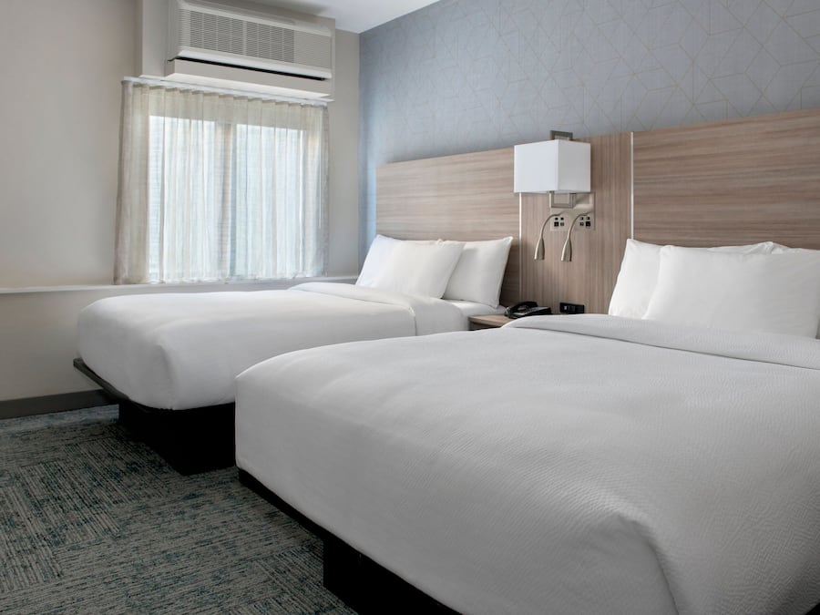 Habitación de hotel con dos camas, una ventana con cortinas, un equipo de aire acondicionado, una mesita de noche con lámpara y un teléfono instalado en la pared.