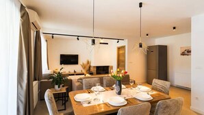A modern dining area with a wooden table set for four, surrounded by upholstered chairs. A flat-screen TV is mounted on the wall above the seating area. - Four Bedroom Holiday Home (Turjaci)