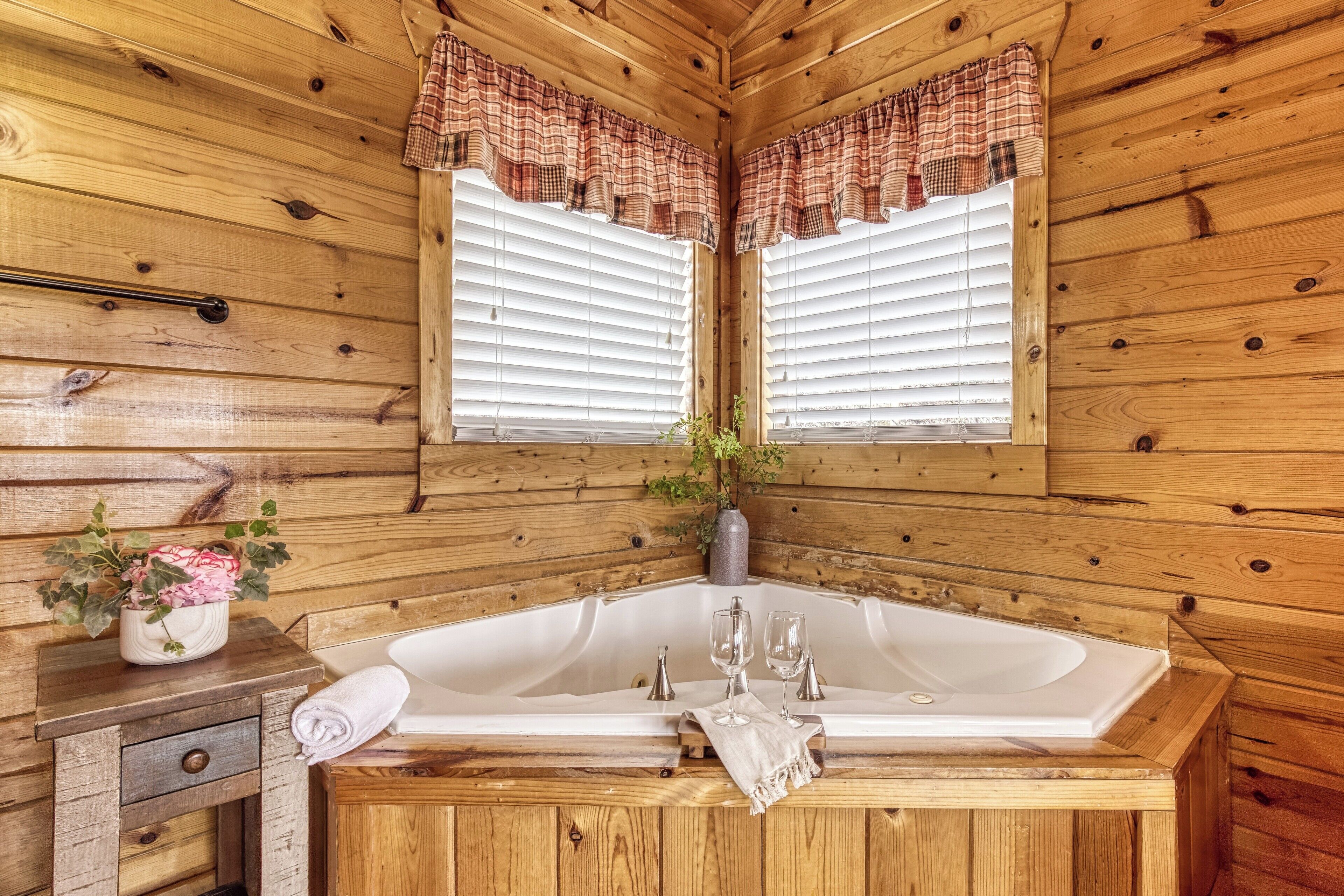A wooden cabin bathroom with a large bathtub, two windows with plaid curtains, a small wooden vanity, and a potted plant.