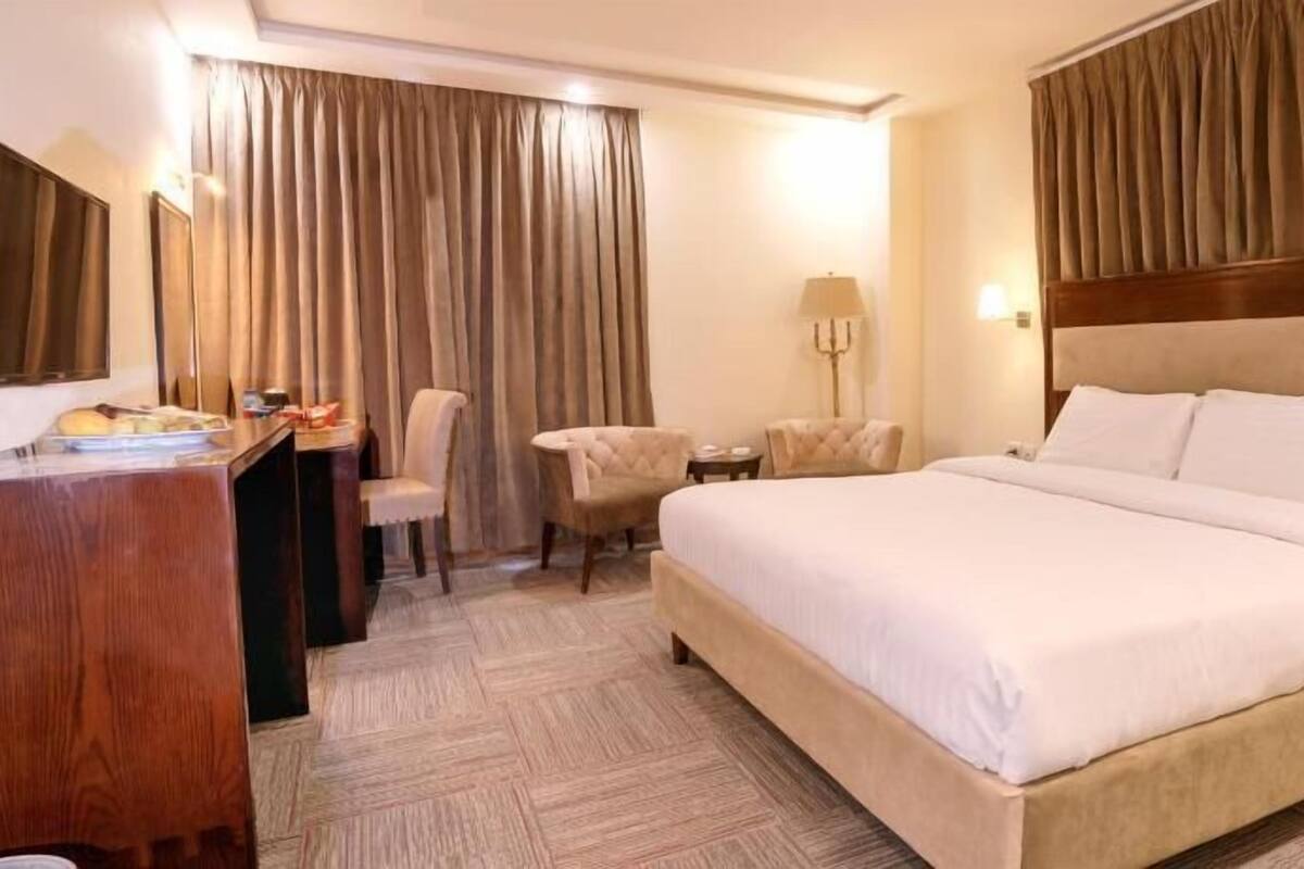 A hotel room with a large bed, a desk, two chairs, a lamp, and curtains.