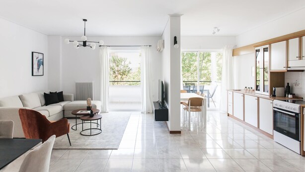 En moderne stue med sofa, sofabord og fjernsyn, et køkken med skabe og hårde hvidevarer, samt store vinduer med udsigt til grønne områder.