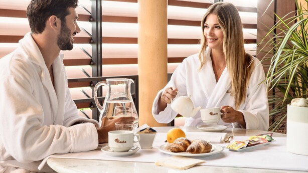 A couple in white robes at a table with a pitcher, cups, pastries, and a plant. - Motel Hotel Piranha (Casalino)
