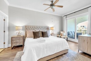 Quarto com uma cama grande, um ventilador de teto, mesa de cabeceira com abajur, uma cadeira e vista para o exterior.