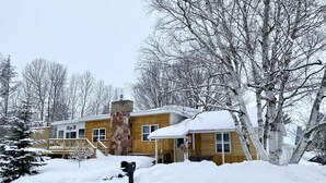 A two-story wooden house with a stone chimney, surrounded by snow-covered trees and a snow-covered deck. - Snowmobile Friendly! Ample Parking-only 1 block to trail! Clean w cozy Fireplace (AuTrain)