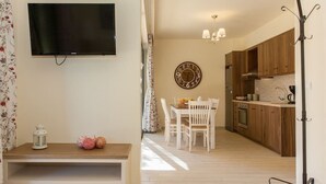 A modern kitchen with wooden cabinets, a dining area with white chairs, a wall-mounted clock, and a flat-screen TV. - Olive Tree Farm (Rethymno)