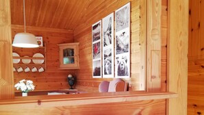 Interior de una cabaña de madera con mostrador de madera, estante con platos, collage de fotografías en la pared y cesta de frutas sobre una mesa.