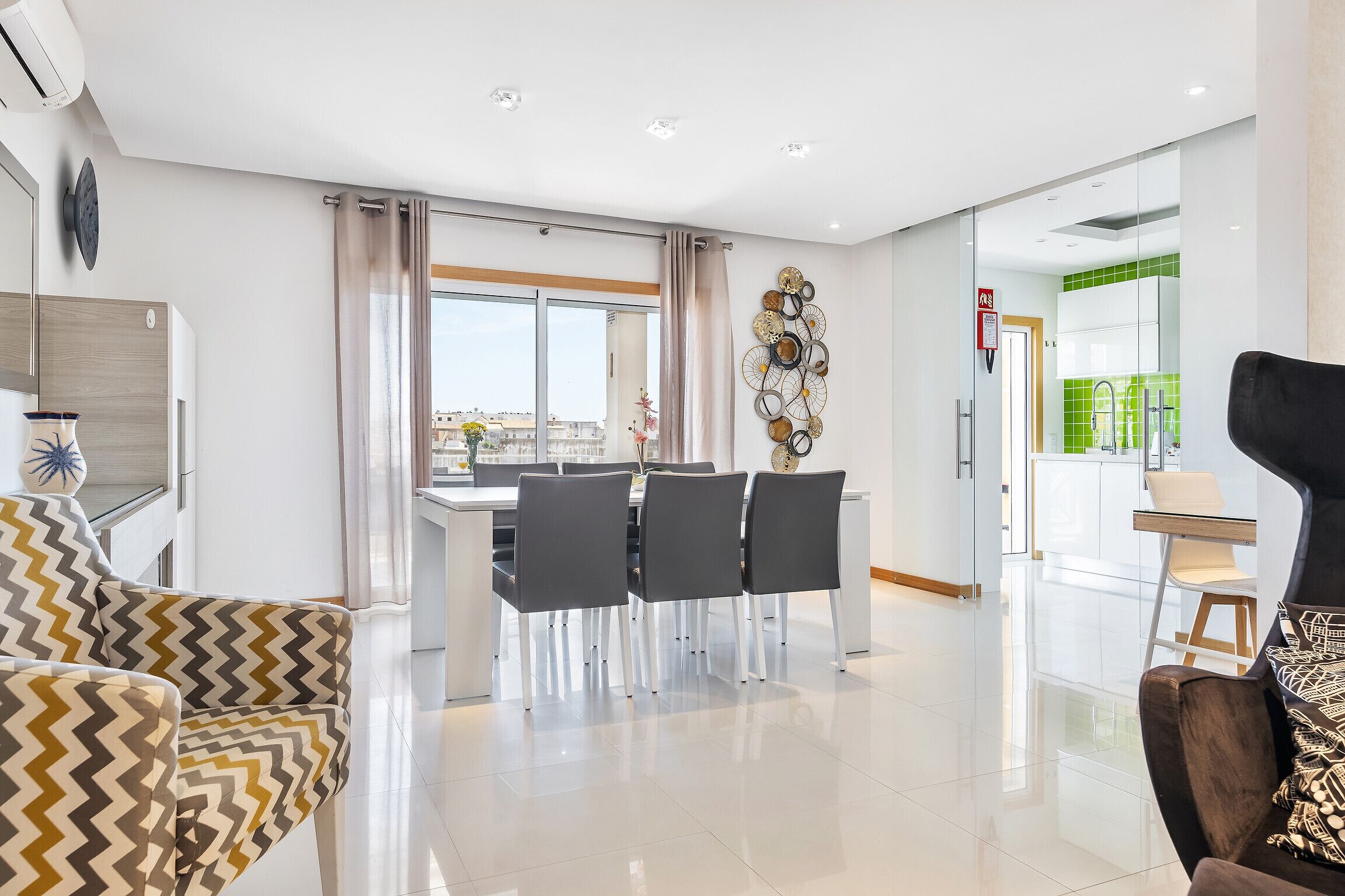 A modern living room with a dining area, a black armchair, and a view of the city through large windows.