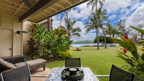 A covered outdoor seating area with a table, chairs, and a view of a grassy landscape with palm trees and a body of water. - Lae Nani 312, Oceanfront, AC,Tropical Bliss, Steps to Beach and Town (Kapaa)