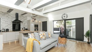 A modern kitchen with a built-in oven, a dining area with a grey sofa and patterned cushions, and a large window with a view of greenery. - The Broadway Beach Homes Airbnb With Hot tub (Herne Bay)