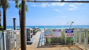 Acceso a la playa con palmeras, una pasarela de madera y varios carteles de advertencia.