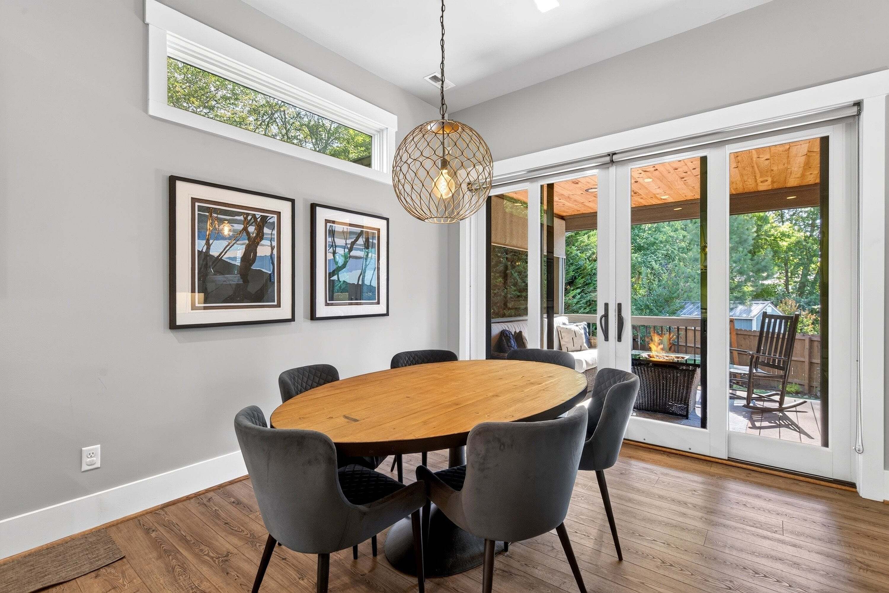 A dining area with a round wooden table, six chairs, two framed pictures, and a hanging light fixture.