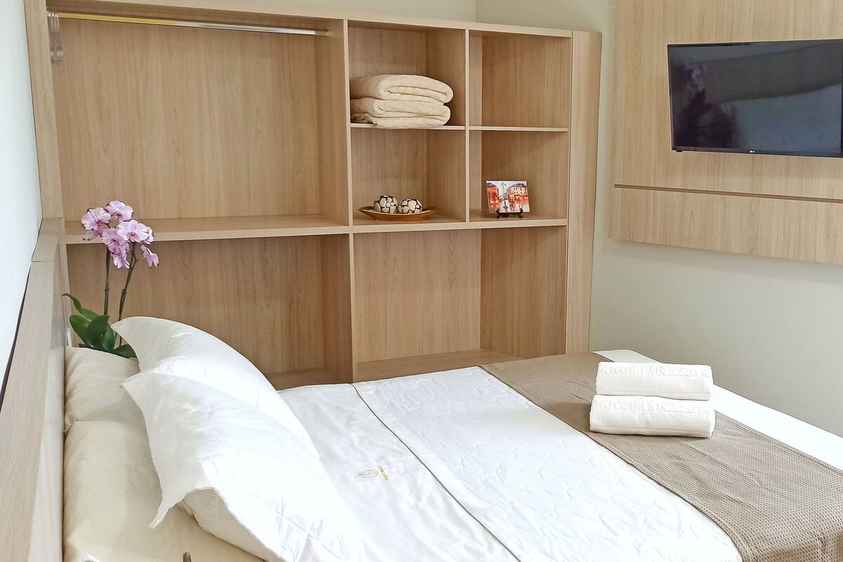 Ein ordentlich eingerichtetes Schlafzimmer mit einem Bett, einem an der Wand montierten Fernseher und einem Holzschrank mit Regalen.
