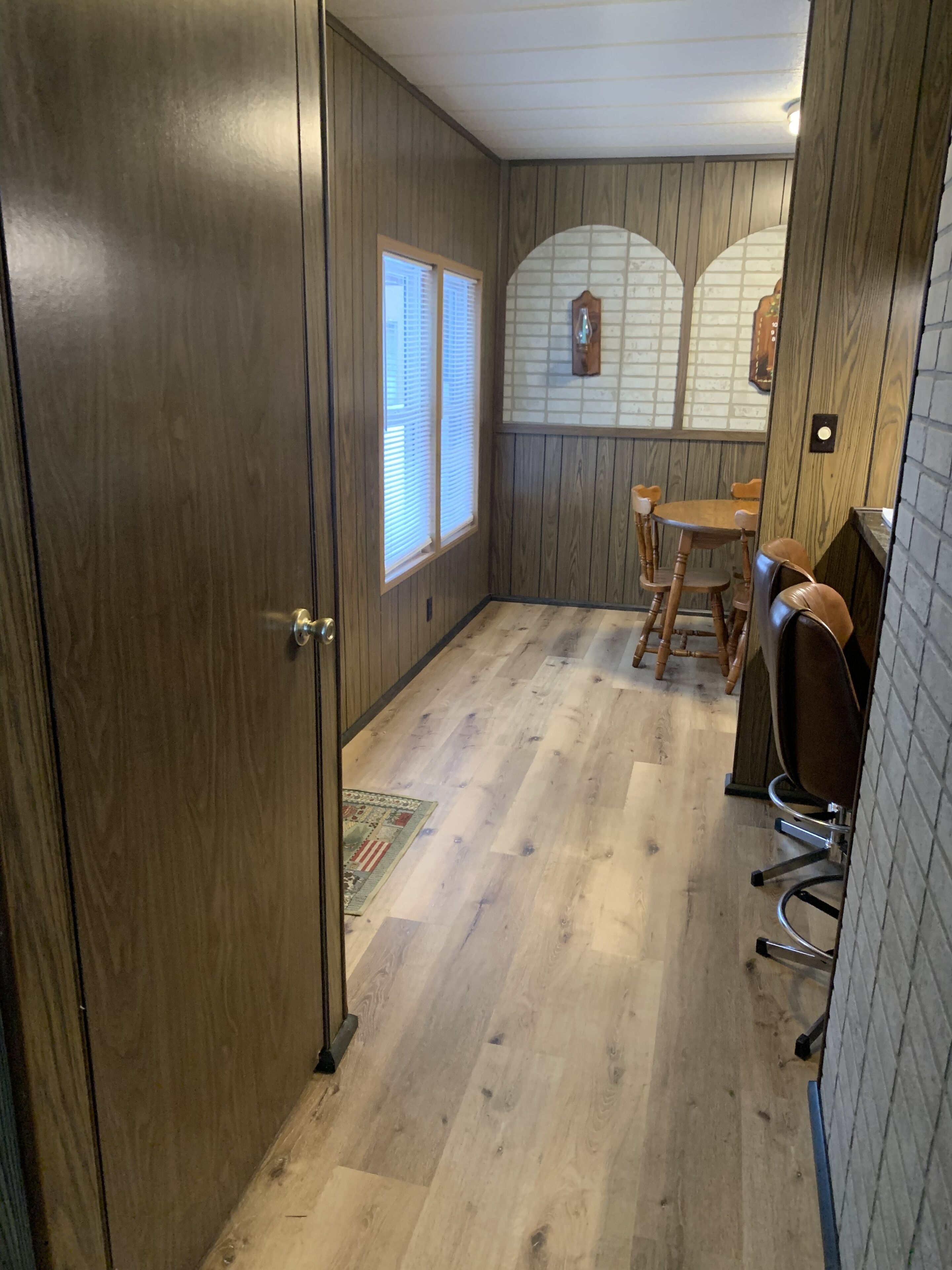 A room with wooden walls, a wooden door, a window with blinds, a wooden table with chairs, and a rug on the floor.