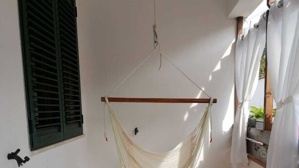 A hammock chair suspended from a wooden beam, a patterned rug on the floor, and a window with green shutters. - Spacious and lovely house (Orosei)