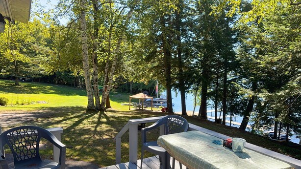 A wooden deck with a table and chairs, overlooking a lake with trees and a gazebo in the background. - Cozy Messalonskee Lake Cabin – Dock, Kayaks & Family‑Friendly, Sleeps 5 (Sidney)