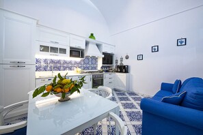 A compact kitchen with white cabinets, a blue and white tiled backsplash, a dining table with a bowl of fruit, and a blue sofa.