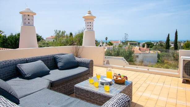 Une terrasse avec un canapé en osier, une table avec des boissons et une vue sur la mer et les bâtiments.