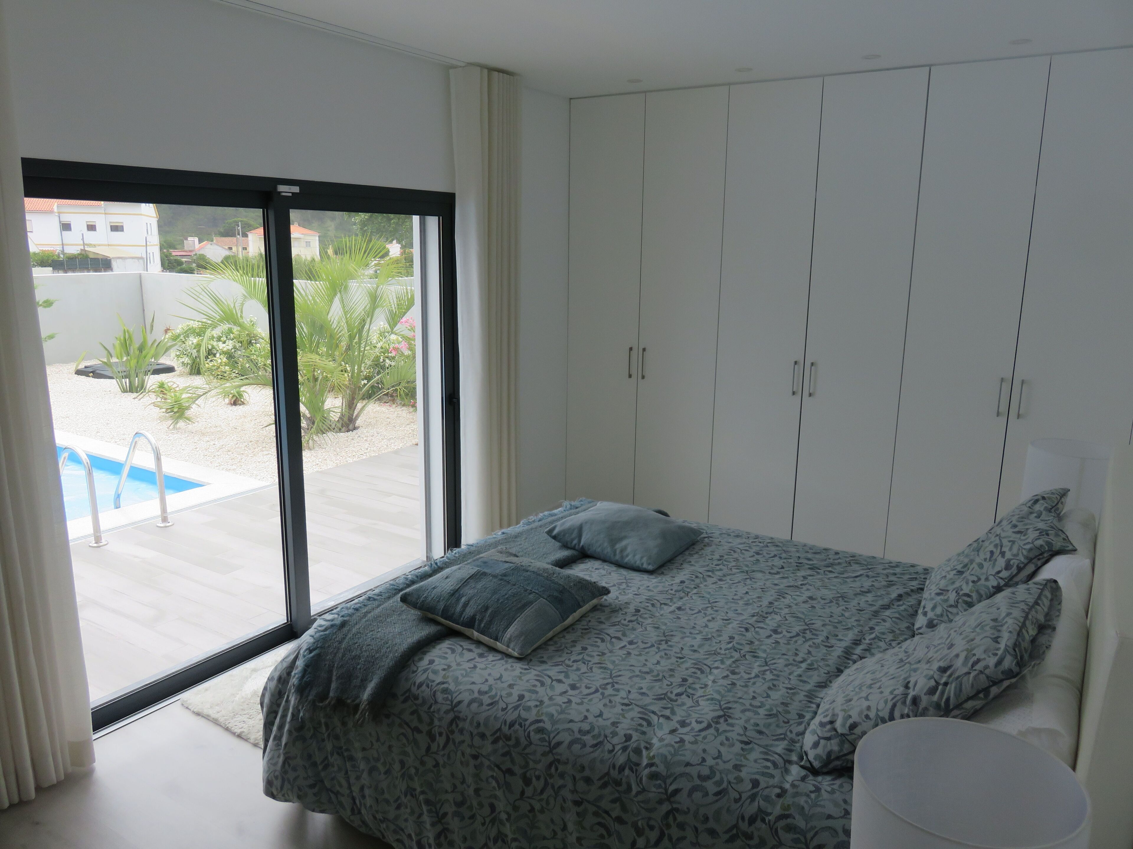 A bedroom with a bed, a white wardrobe, and a view of a swimming pool and greenery outside.