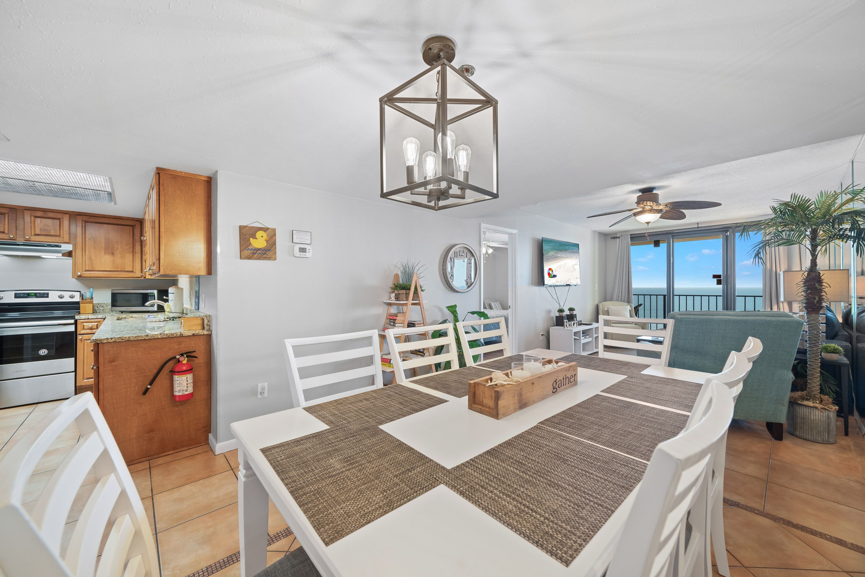 A modern kitchen with wooden cabinets, a microwave, and a gas stove. There is a dining area with a white table and chairs, and a view of the ocean through large windows.