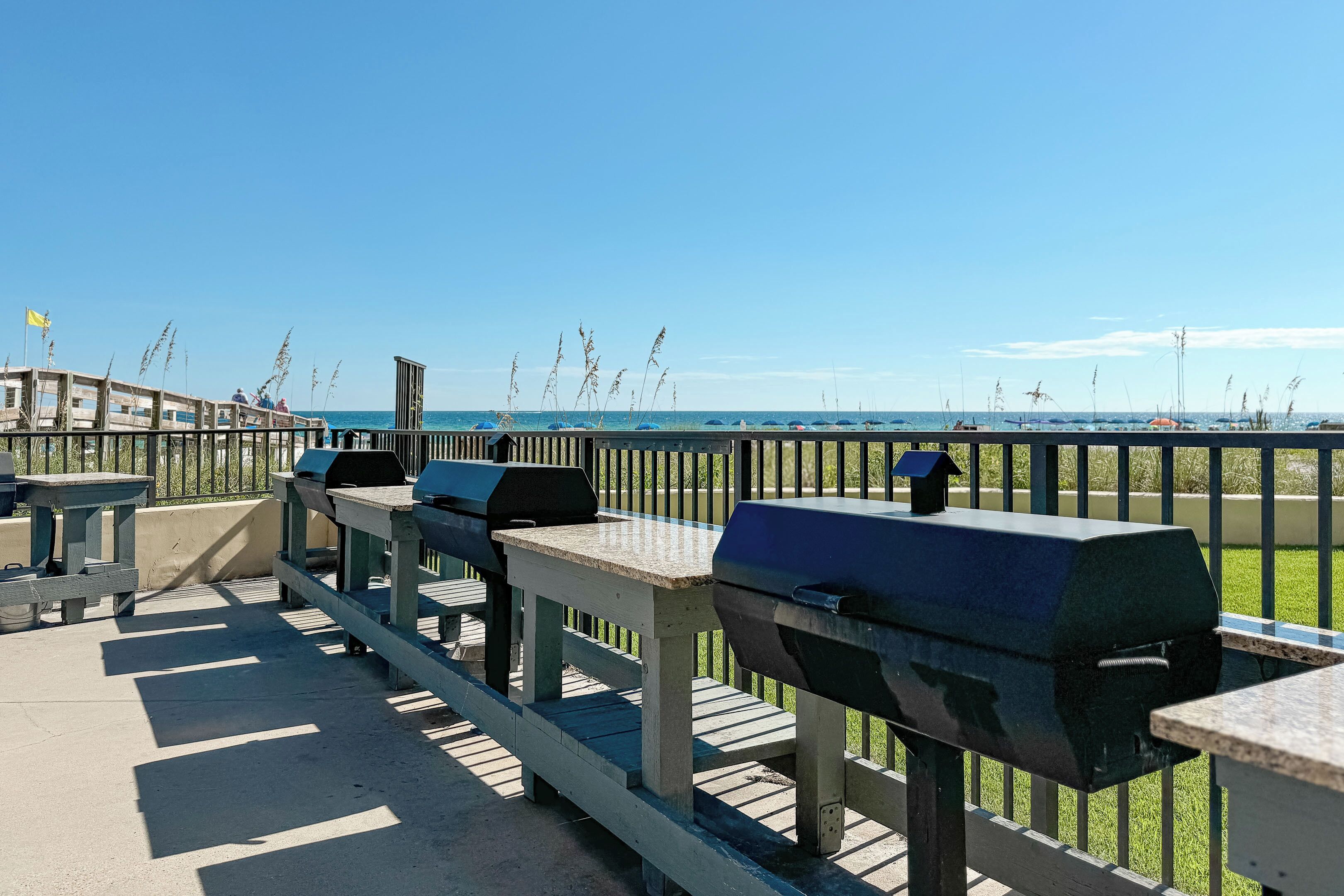 Terraza exterior con mesas de picnic y parrillas, con vista a una playa bajo un cielo despejado.