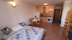 Una camera compatta con un letto, comodino e una piccola zona pranzo con tavolo e sedie. Adiacente, una kitchenette con piano cottura, forno e lavello.