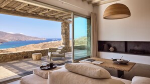 A modern living room with a large glass door leading to a terrace overlooking a coastal landscape. - Villa Amira NEW Luxury Property in Ftelia with Amazing Sea views & Heated pool (Mikonos)