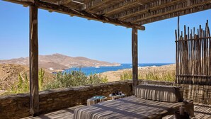 A wooden deck with a stone wall, a cushioned seating area, and a view of the sea and mountains. - Villa Amira NEW Luxury Property in Ftelia with Amazing Sea views & Heated pool (Mikonos)