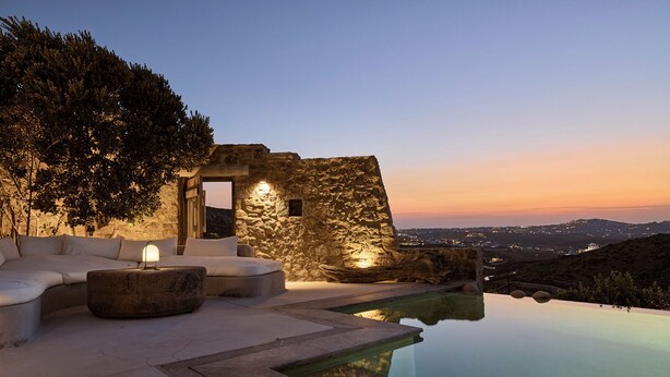 A luxurious outdoor seating area with a stone wall, a pool, and a view of the landscape. - Villa Amira NEW Luxury Property in Ftelia with Amazing Sea views & Heated pool (Mikonos)