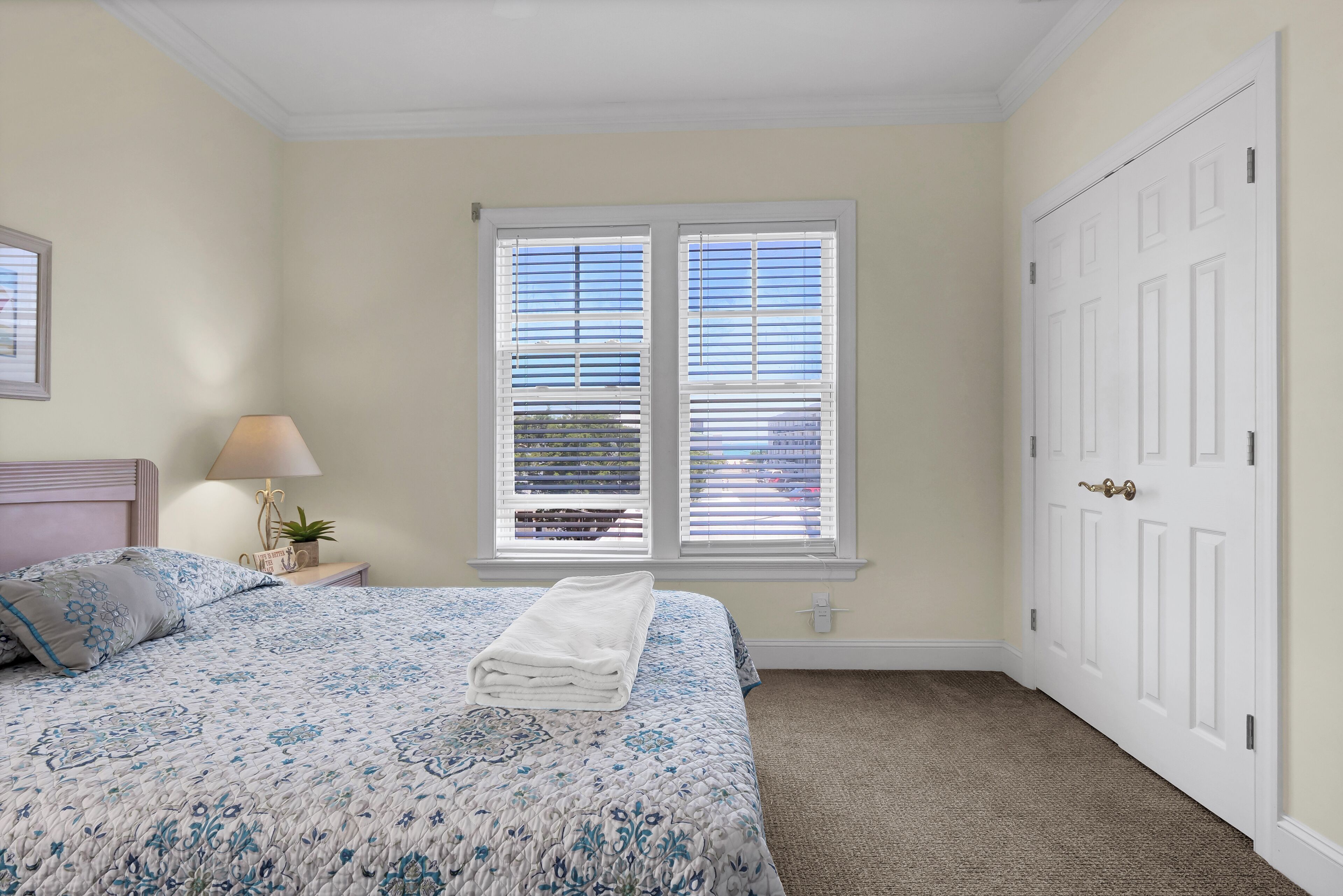 Un dormitorio con cama, dos ventanas con persianas, una mesita de noche con lámpara y una puerta.
