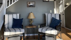 A staircase with white railings leading to an upper level. Two chairs with patterned cushions are placed on a patterned rug in the lower area. A lamp with a wooden base is on a small table between the chairs.