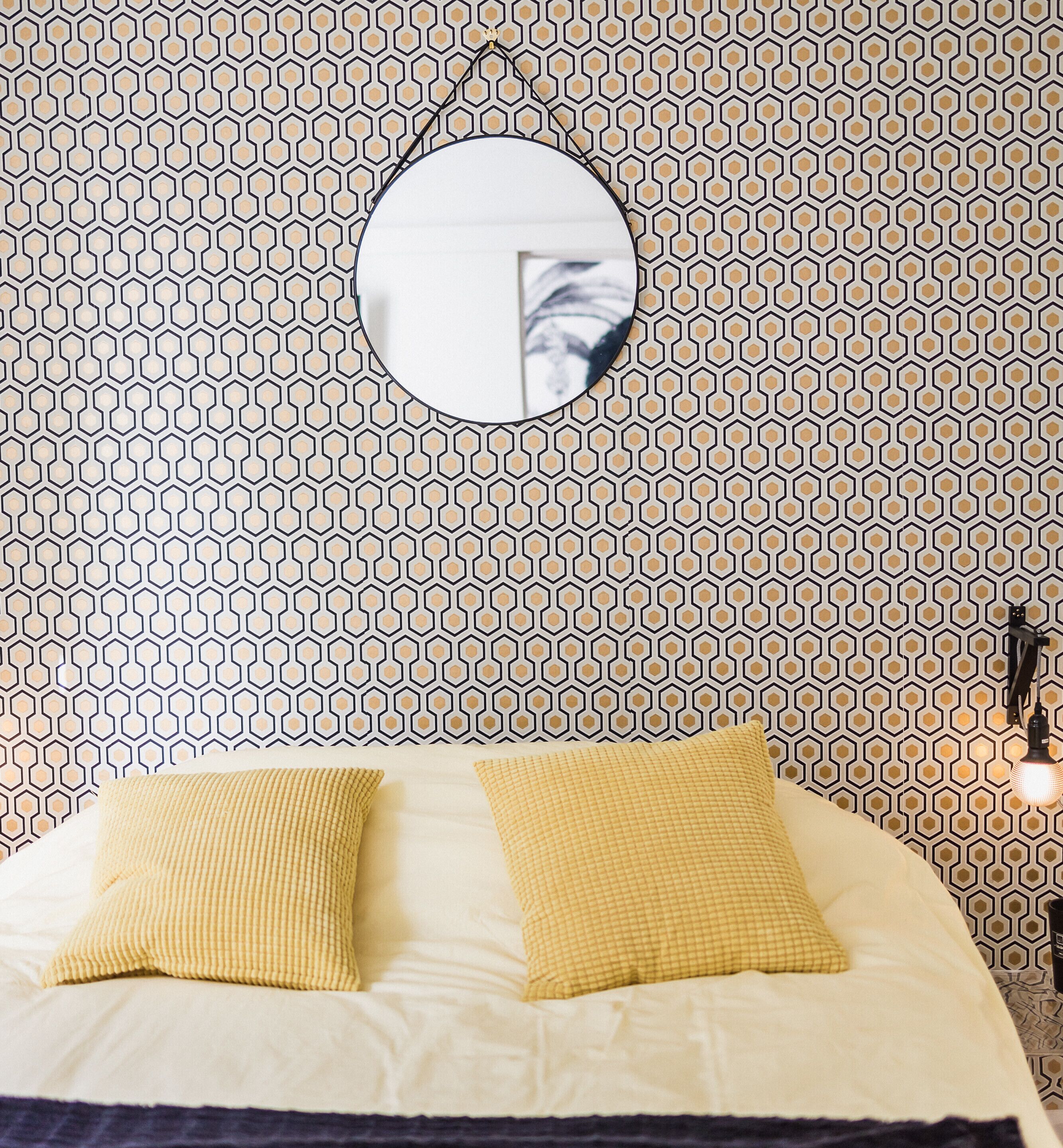 A bedroom with a patterned wallpaper, a round mirror, and two yellow pillows on a bed.