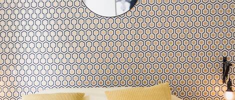 A bedroom with a patterned wallpaper, a round mirror, and two yellow pillows on a bed.