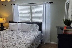 A bedroom with a bed, white curtains, a bedside table with a lamp, and a small plant. - 5388 Leaf Back · Lacinda`s Cozy Home (Beckett Ridge)