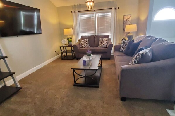 A living room with a sofa set, a coffee table, a TV mounted on the wall, and a window with curtains. - 5388 Leaf Back · Lacinda`s Cozy Home (Beckett Ridge)