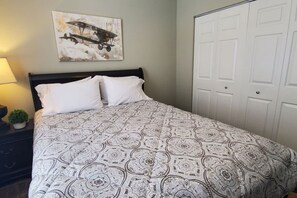 Un dormitorio con cama, almohadas, una mesita de noche con lámpara, una planta pequeña y una puerta.