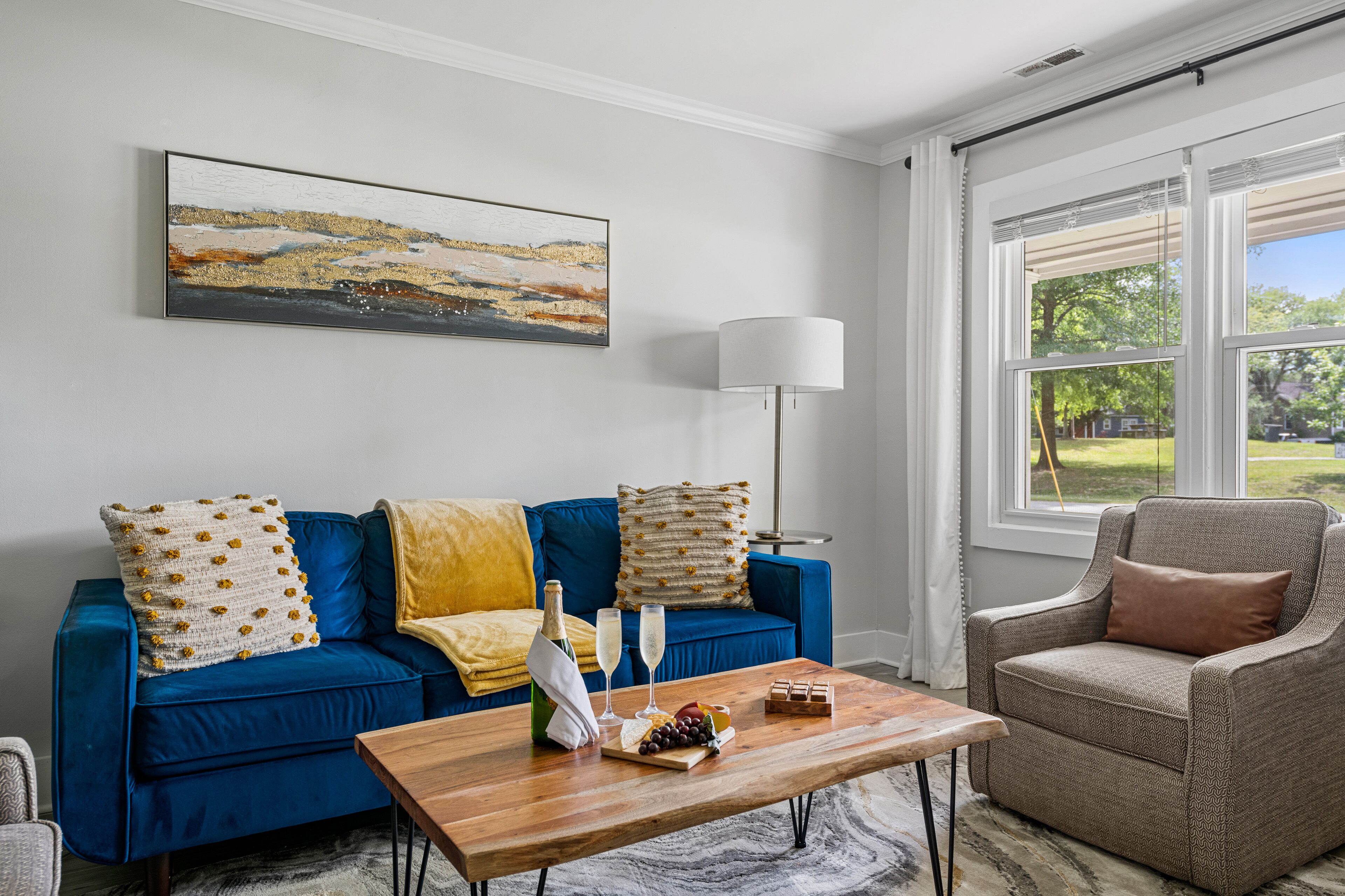 Un salon avec un canapé bleu, un fauteuil marron, une table basse en bois sur laquelle se trouvent une bouteille, des verres et des en-cas, ainsi qu’un tableau accroché au mur.