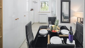 Un coin repas avec une table noire dressée pour deux personnes, des assiettes blanches et une bouteille de vin. On y trouve également un panier de fruits contenant de l’ananas et d’autres fruits, et une fenêtre avec des stores en arrière-plan.