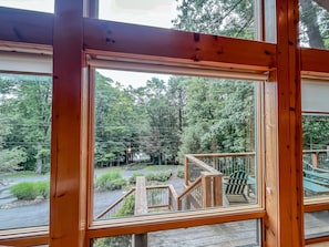 A wooden-framed window with a view of a deck, outdoor seating, and surrounding greenery.